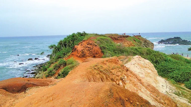 A scenic view of the Ussangoda Ramayana Site in Ussangoda National Park, Sri Lanka. Red soil cliffs rise above the turquoise waters of the Indian Ocean, with lush vegetation growing on the slopes.